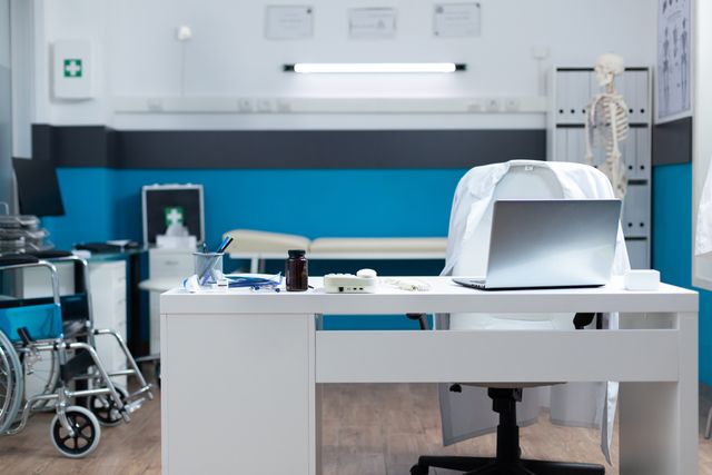 Medical consultation room with a white desk, laptop, wheelchair and examination equipment against a blue accent wall.