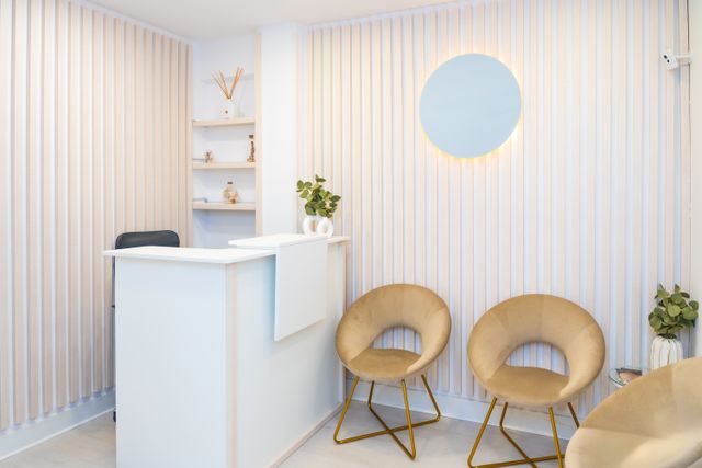 Compact reception with pale vertical wall slats, a white counter, rounded gold-toned chairs and a circular backlit wall feature.
