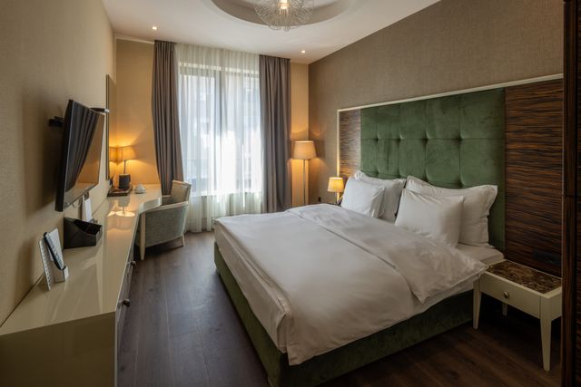 Hotel guestroom with a green upholstered headboard, white bedding, writing desk and layered window curtains.