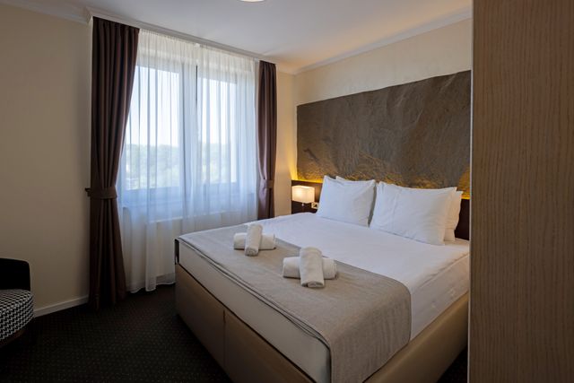 Hotel bed against a dark textured headboard wall beside soft curtains and warm bedside lighting.