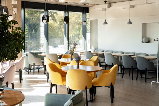 Bright cafe with yellow and grey upholstered chairs, round tables, pendant lights and large street-facing windows.
