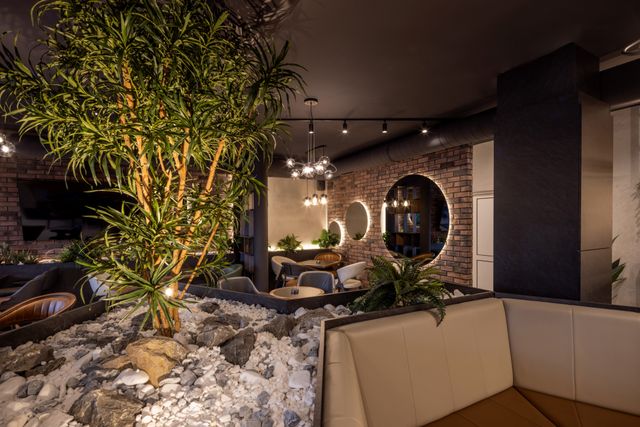 Restaurant seating arranged around an illuminated indoor tree and stone planting bed, with warm lighting and brick wall accents.