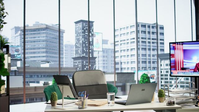 Light executive desk with a laptop and task chair beside full-height city-facing windows.