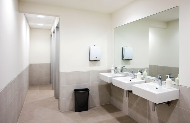 Shared washroom with three white wall-mounted basins, a wide mirror and pale grey tiled surfaces.