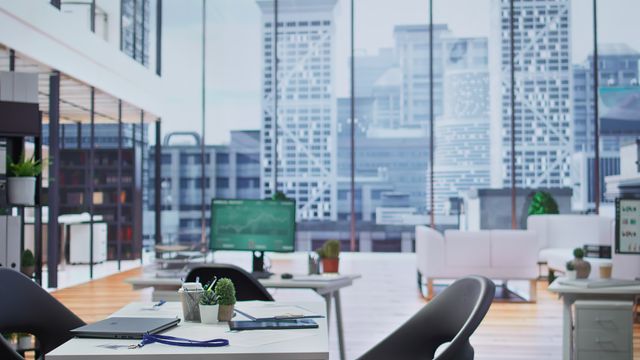 White office desks with dark chairs, desktop monitors and small plants, with lounge seating in the background.