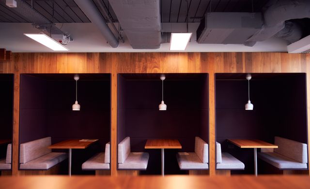 A row of high-sided upholstered seating booths with small tables and pendant lights beneath an exposed-services ceiling.