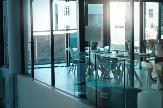 Daylit meeting room viewed through a glass partition, with a long white table, chairs, laptops and a presentation board.