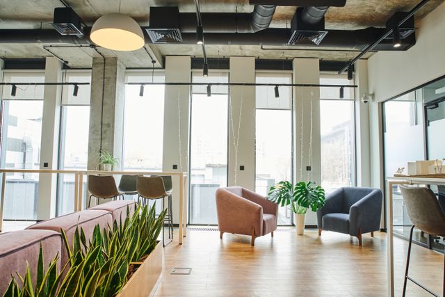 Workplace lounge with upholstered armchairs, a sofa, indoor planting and high tables beneath an exposed-services ceiling.