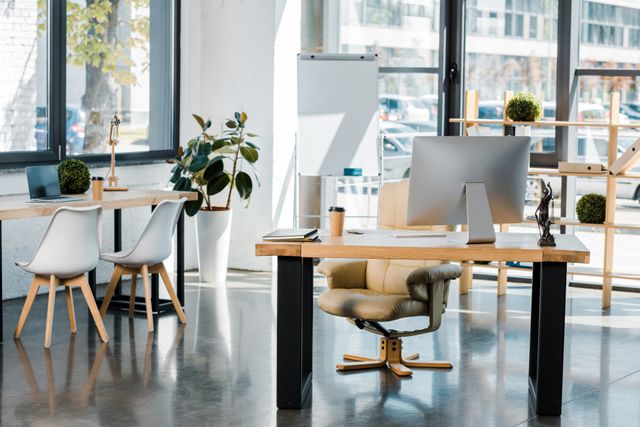Timber office desks with a desktop monitor, open shelving, plants and a white presentation board beside large windows.