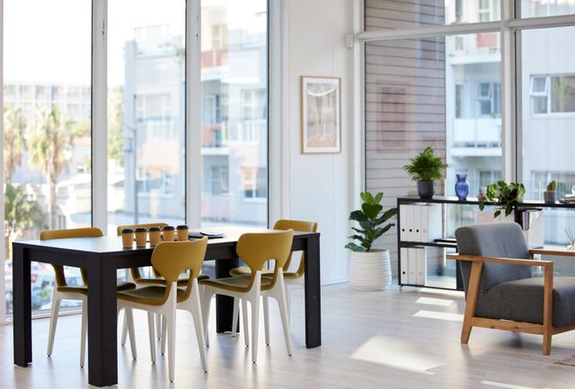 Shared table with mustard-coloured chairs beside a grey lounge chair, low shelving and sunlit windows.