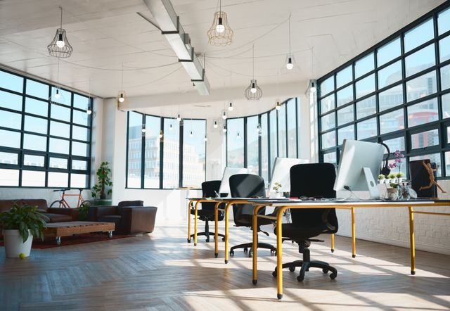 Open office with a row of slim desks and task chairs, large black-framed windows, pendant bulbs and a lounge area.