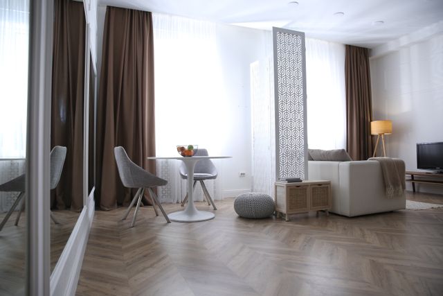 Open living and dining area with parquet flooring, tall curtains and neutral seating.