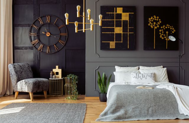 Grey bedroom with wall mouldings, upholstered bed, gold-tone lighting and a floral armchair.