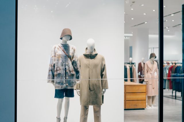 Two mannequins displayed behind a large shop window with a clear view of timber fixtures and apparel rails inside.