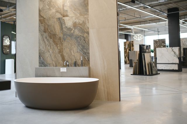 Freestanding oval bath in front of a tall stone-finish display with sample panels and showroom aisles beyond.