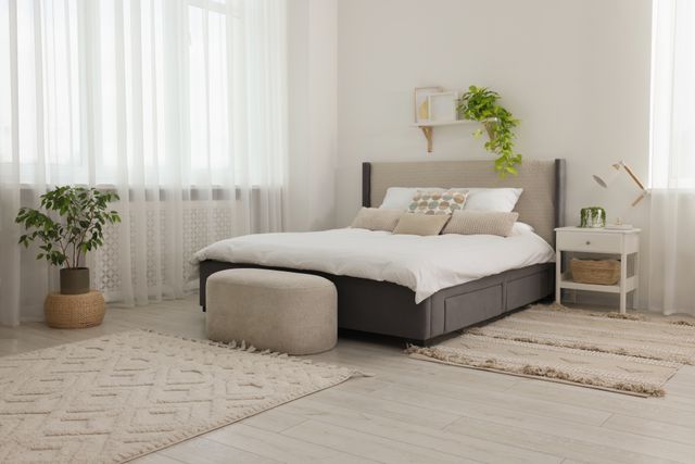 A grey upholstered bed and matching ottoman sit beside a window in a softly lit neutral bedroom.