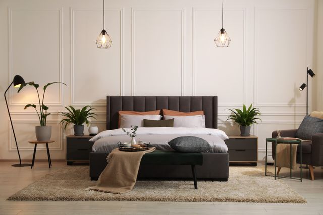A grey bed and armchair are framed by white wall molding, pendant lights and indoor plants.