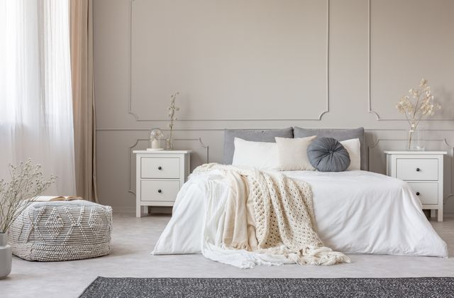 A grey headboard, white bedside tables and a chunky knitted throw furnish a light neutral bedroom.