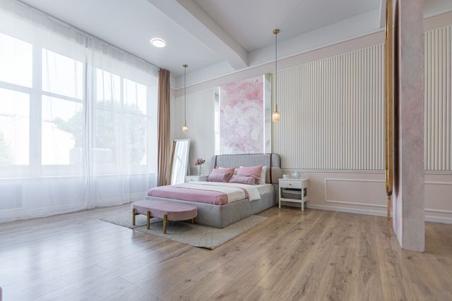 A bed with pink accents stands beneath pendant lights beside tall windows and finely textured wall panels.