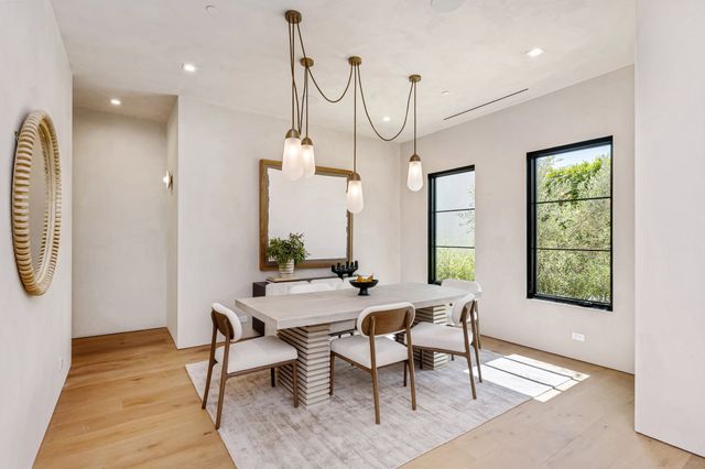 A dining table with upholstered chairs sits beneath pendant lights between large windows and warm wood flooring.