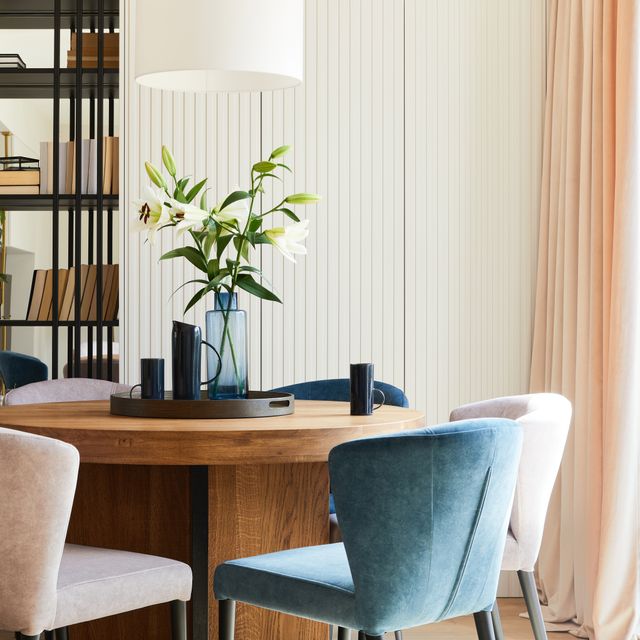 Velvet dining chairs surround a rounded timber table with decorative vases near softly colored curtains.