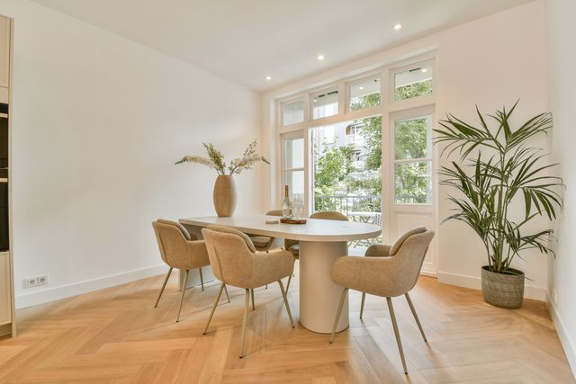 A round dining table and curved woven chairs sit beside a large window and a tall indoor plant.