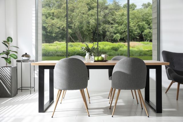A wood dining table and grey upholstered chairs face wide windows overlooking green planting.