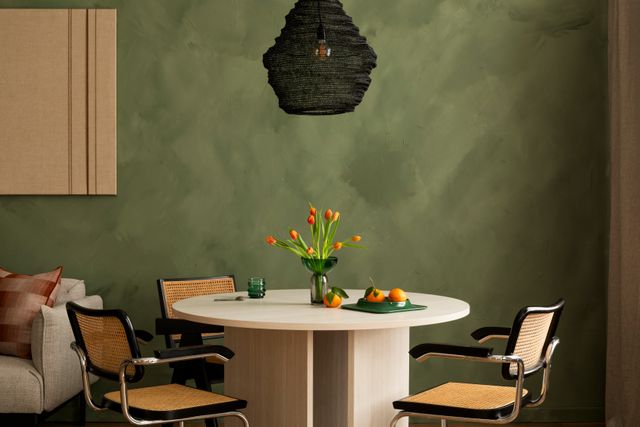 A round pale table and woven chairs sit against a textured green wall beneath a dark pendant.