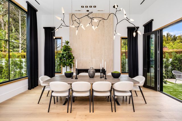 A long dining table with pale chairs sits beneath a branching chandelier between tall garden-facing windows.
