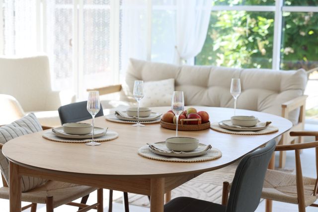 An oval dining table is set with plates, cups and glassware in a bright room with soft seating beyond.