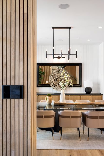 A dining area with upholstered chairs and a chandelier is viewed past timber paneling toward a large mirror.