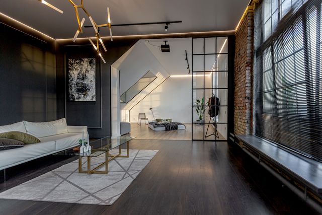 A charcoal living area with pale seating connects to a bright bedroom beyond a glass partition.