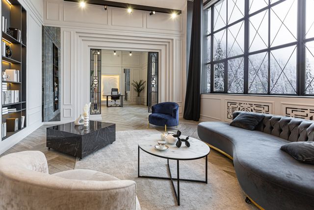 A dark curved sofa and marble tables sit beside tall windows in a room with white paneled walls.