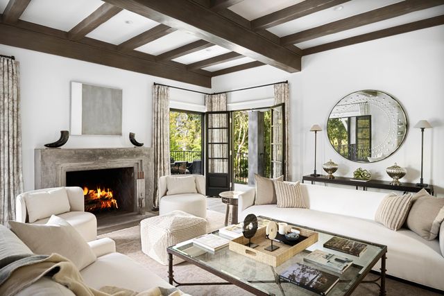 White sofas surround a glass coffee table near a stone fireplace beneath dark exposed ceiling beams.