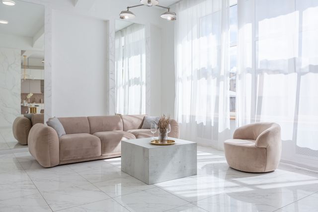 Rounded beige seating surrounds a pale stone table beside tall windows dressed with sheer curtains.