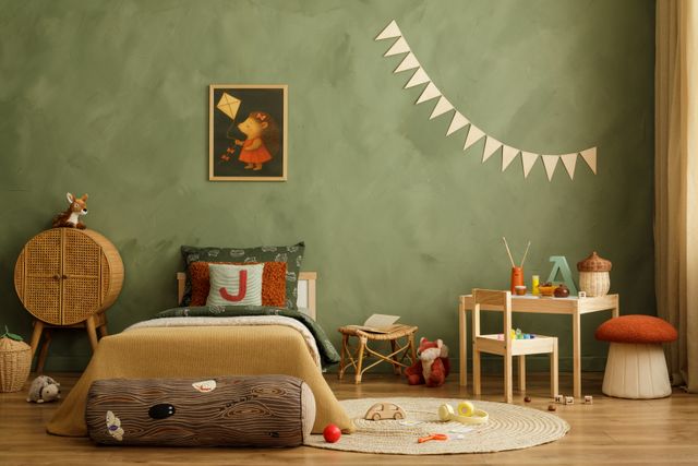 A small bed, woven cabinet and play table are arranged against a green wall with forest-themed details.