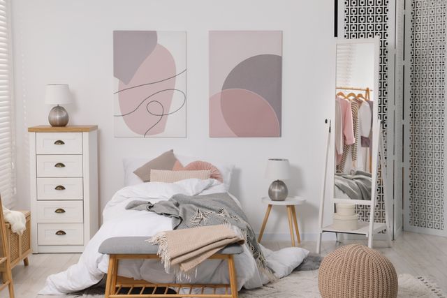 A single bed with layered neutral bedding sits beneath abstract artwork beside drawers and an open clothes rail.