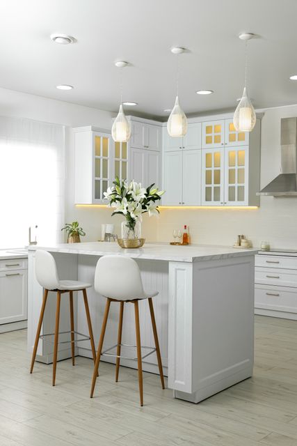 A white kitchen island with timber-legged stools sits beneath pendant lights beside glazed upper cabinets.