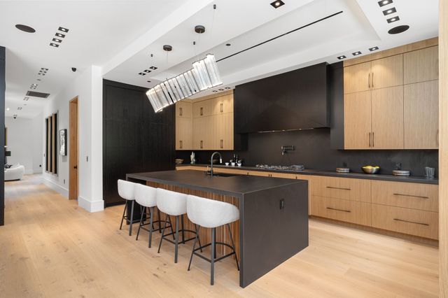 A long dark island with pale bar stools anchors a kitchen lined with timber cabinets and black surfaces.
