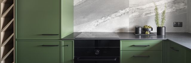 Green cabinet fronts surround a dark worktop and oven beneath a grey stone-look kitchen backsplash.