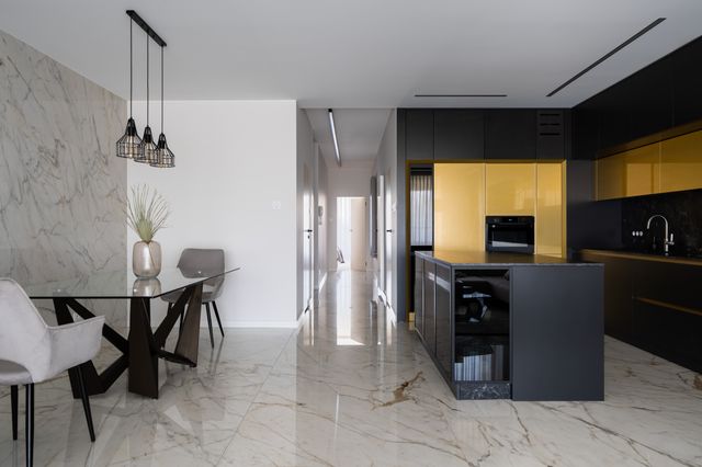 Black and golden cabinetry frame a kitchen island beside a dining table and pale stone-look flooring.