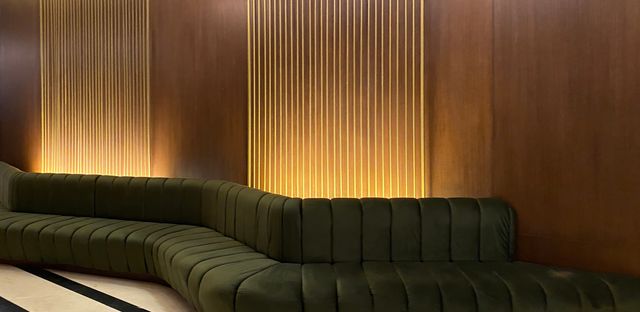 A curved dark-green sofa follows a timber wall accented by slender vertical lines and warm concealed lighting.
