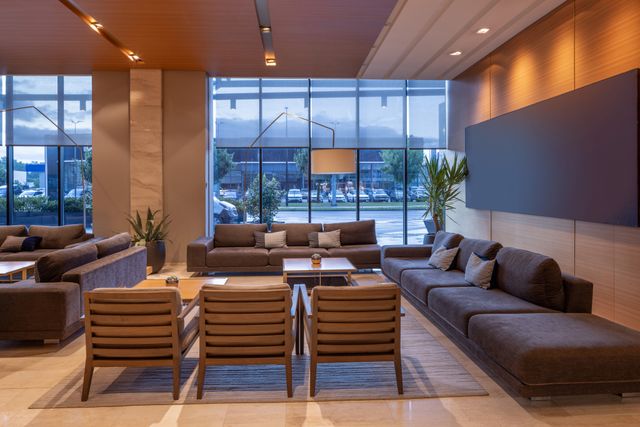 Sofas and timber chairs form a spacious lobby seating area beside tall windows and warm wall lighting.