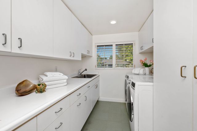 White storage cabinets, laundry appliances and a long worktop line a utility room with a window at the end.
