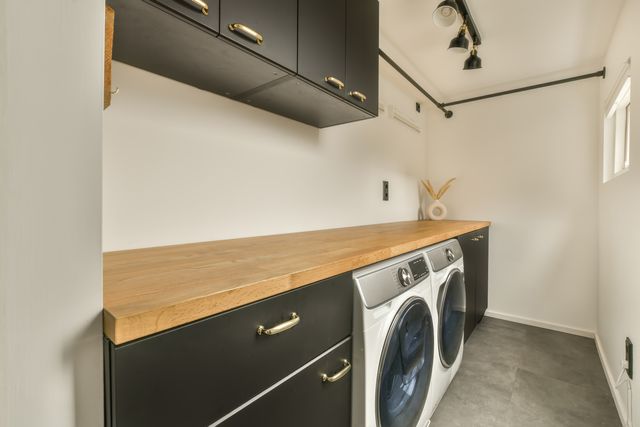 Front-loading laundry appliances sit below a timber worktop and dark storage cabinets with warm metal handles.