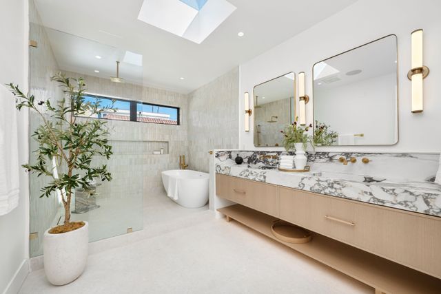 A long timber vanity with patterned stone, rounded mirrors and warm metal fixtures faces a skylit bathing area.
