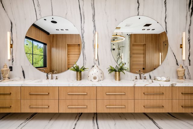 Two round mirrors hang above a long timber vanity with pale veined stone and warm wall lights.