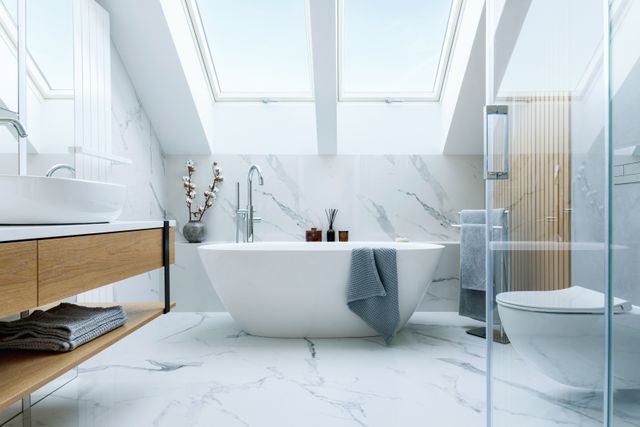 A freestanding white bathtub sits below two roof windows beside timber storage and pale marble-patterned surfaces.