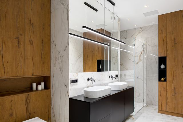 Twin white basins, dark cabinetry and linear pendant lights sit between timber cupboards and a glass shower.