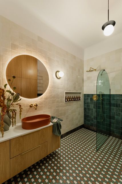 A terracotta basin, circular mirror and green-tiled shower are paired with timber cabinetry and patterned floor tiles.
